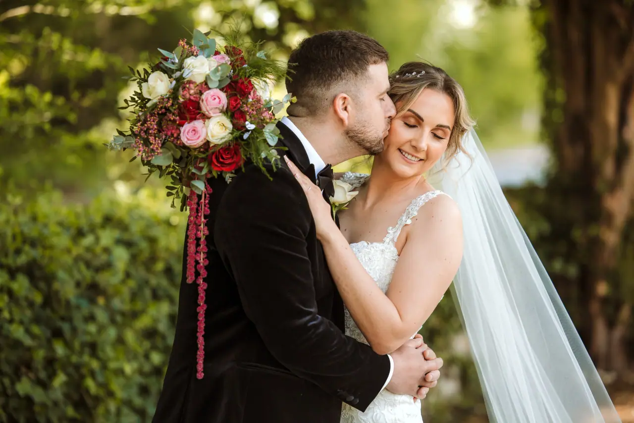 Alex kissing Olivia's cheek as she holds a red and pink rose bouquet