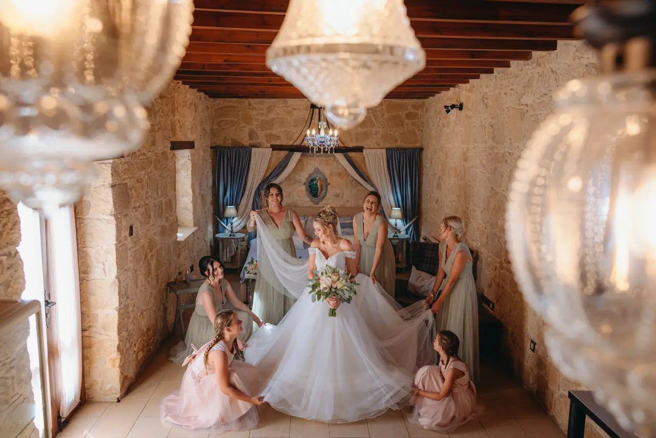 Bride's skirt spread by bridesmaids and flower girls in a stone room under a chandelier, Liopetro