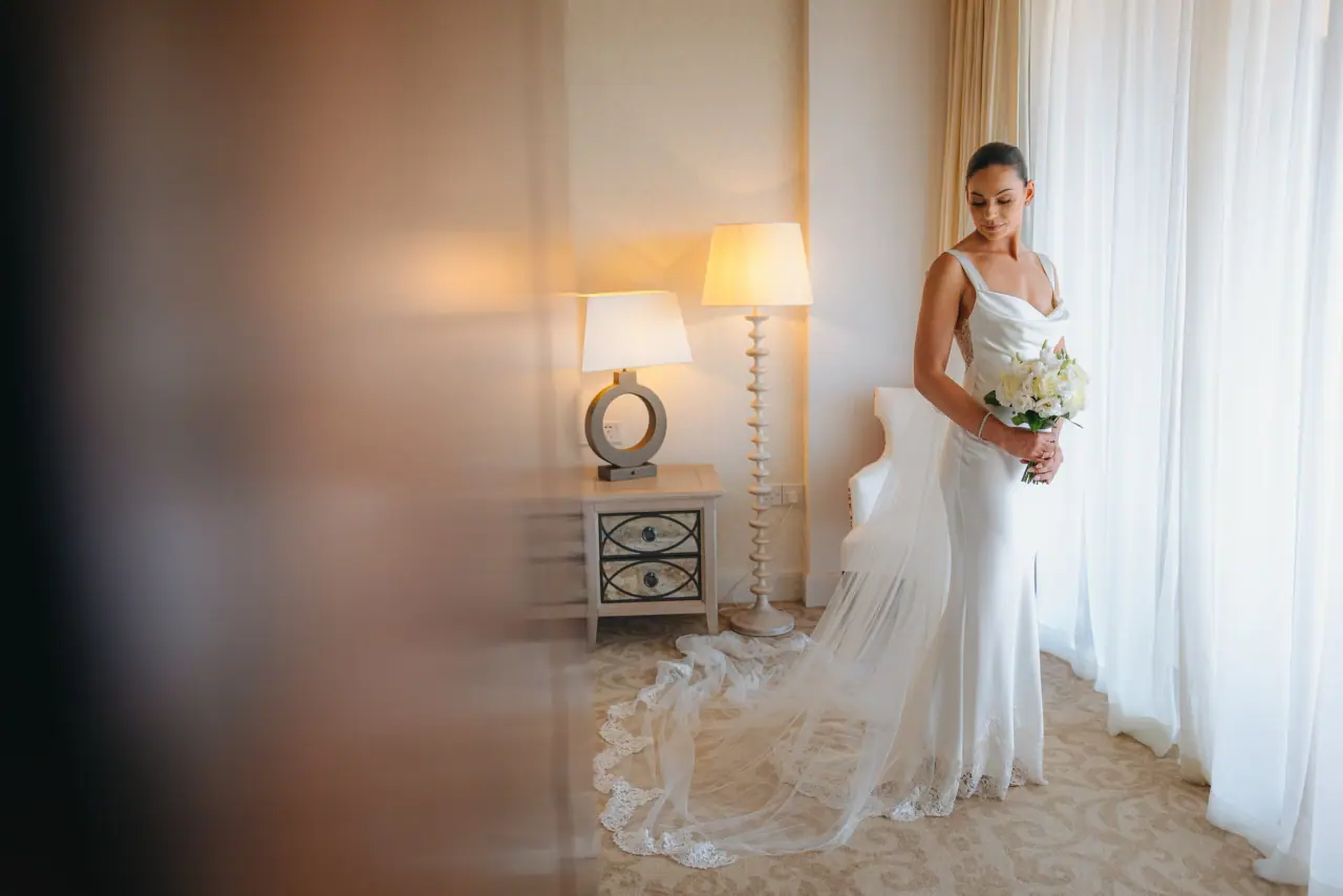 Amy holding her bouquet in a sunlit bedroom, her lace train behind her, Alassos