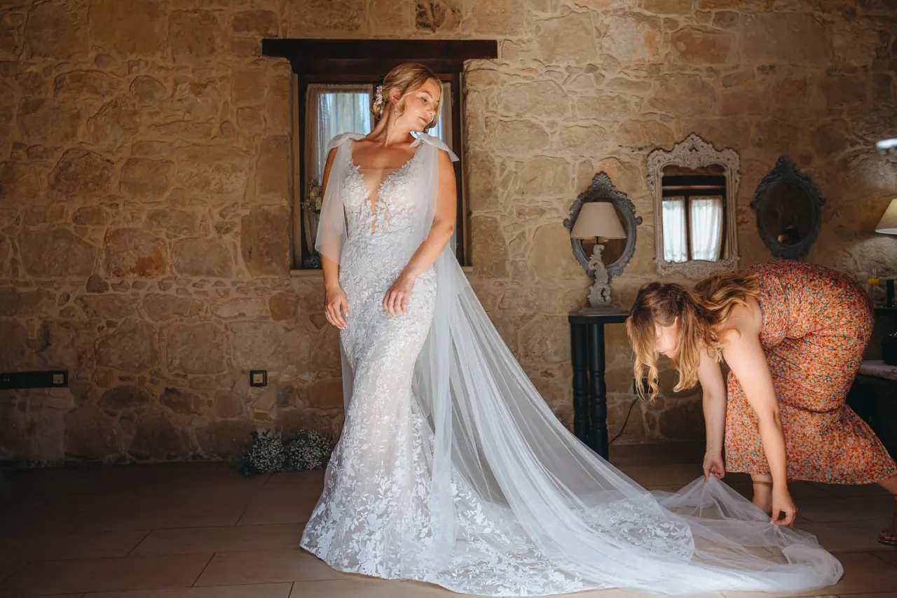 Bride in a long veil as a guest adjusts her train in a stone room at Liopetro