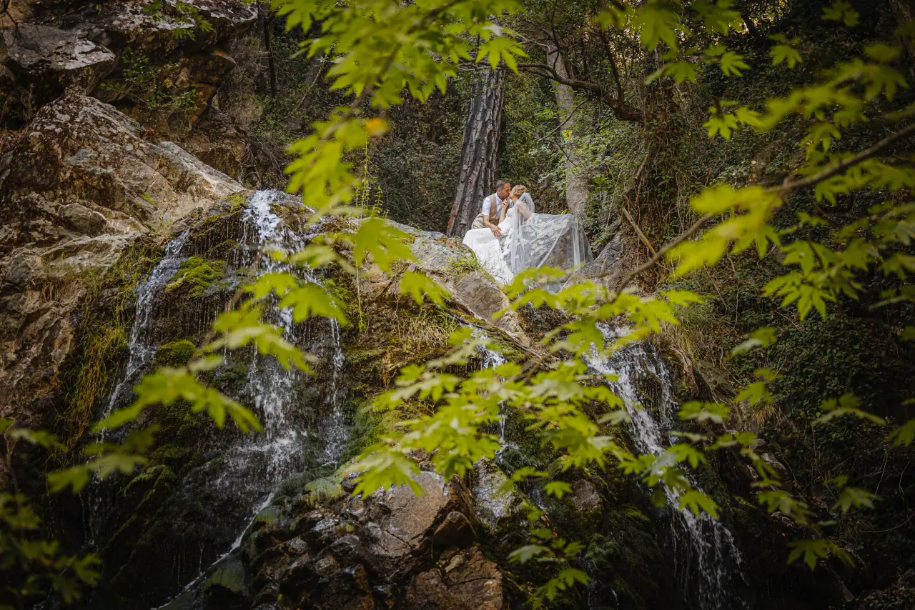 Carrie and Chris on mossy rocks beside a forest waterfall, framed by green leaves