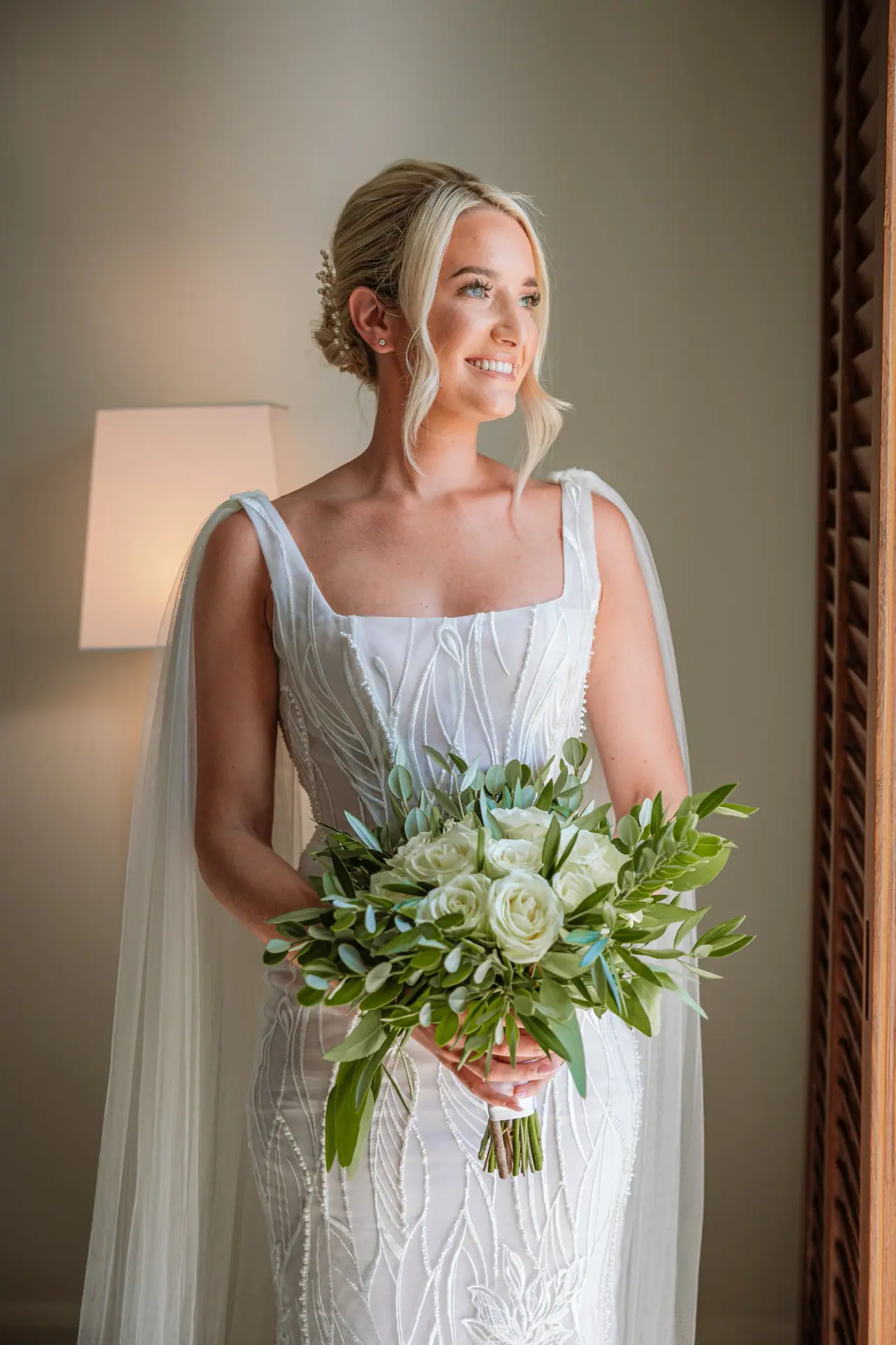 Charlotte smiling with a white and green bouquet by a window at Alassos