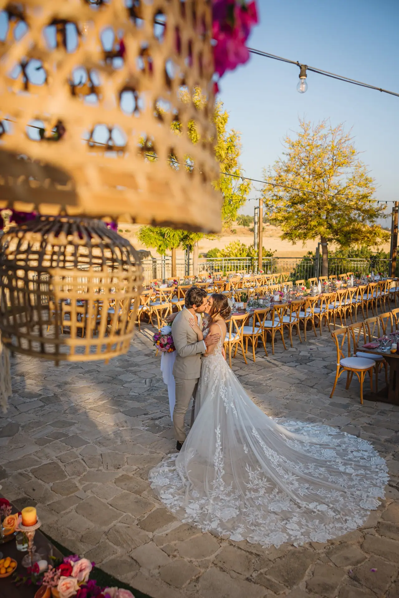 Bride with a long lace train in a stone courtyard beside a tall wicker lantern, Liopetro