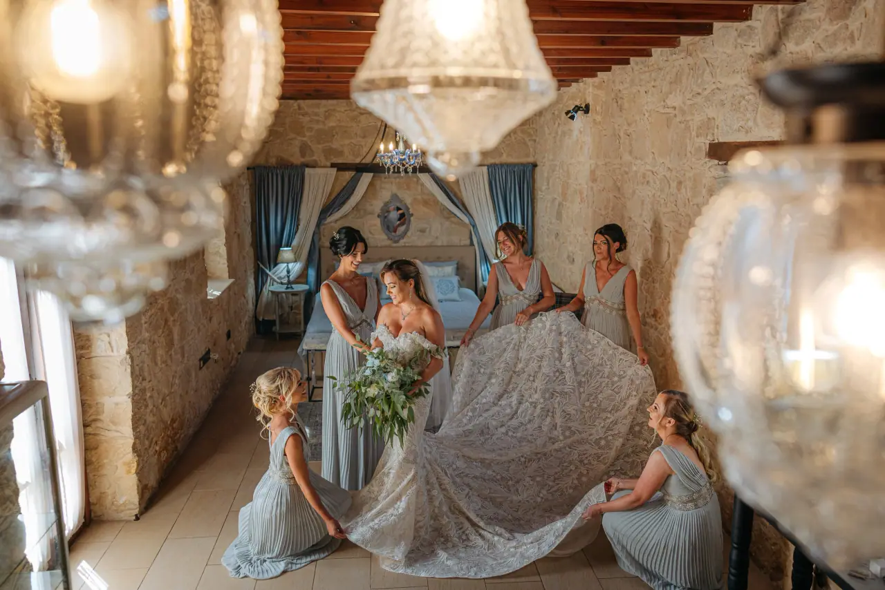 Bridesmaids spreading Annie's lace train in a stone room beneath a chandelier