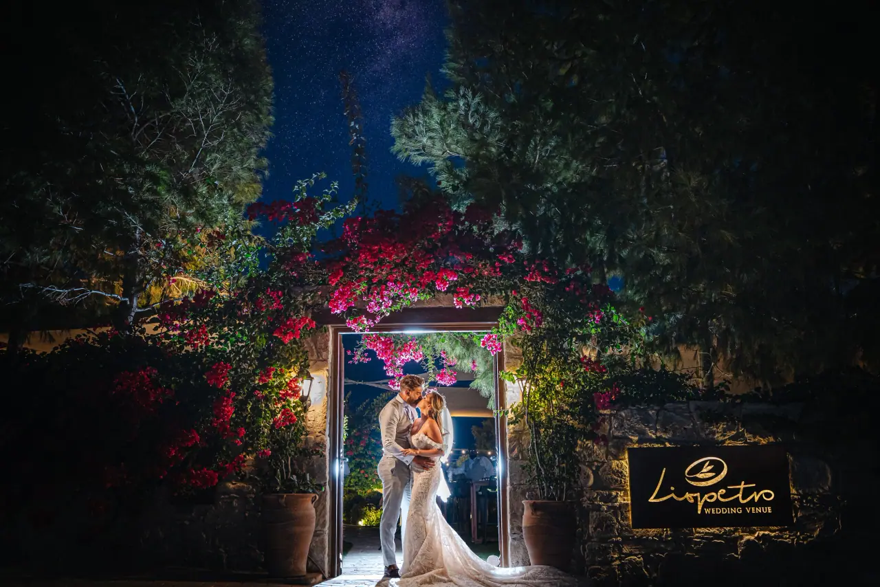 Annie and Iain kissing in a lit, bougainvillea-framed doorway at night, Liopetro