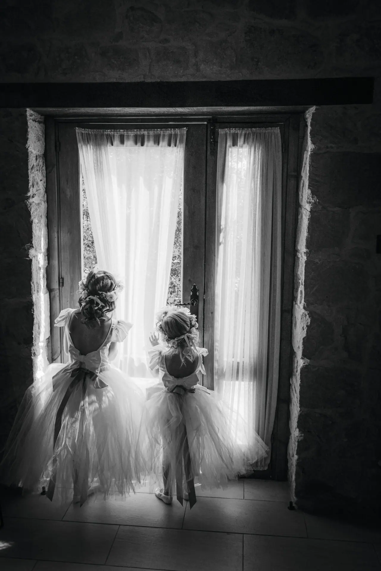 Black-and-white photo of two young bridesmaids in tulle dresses looking out of a window