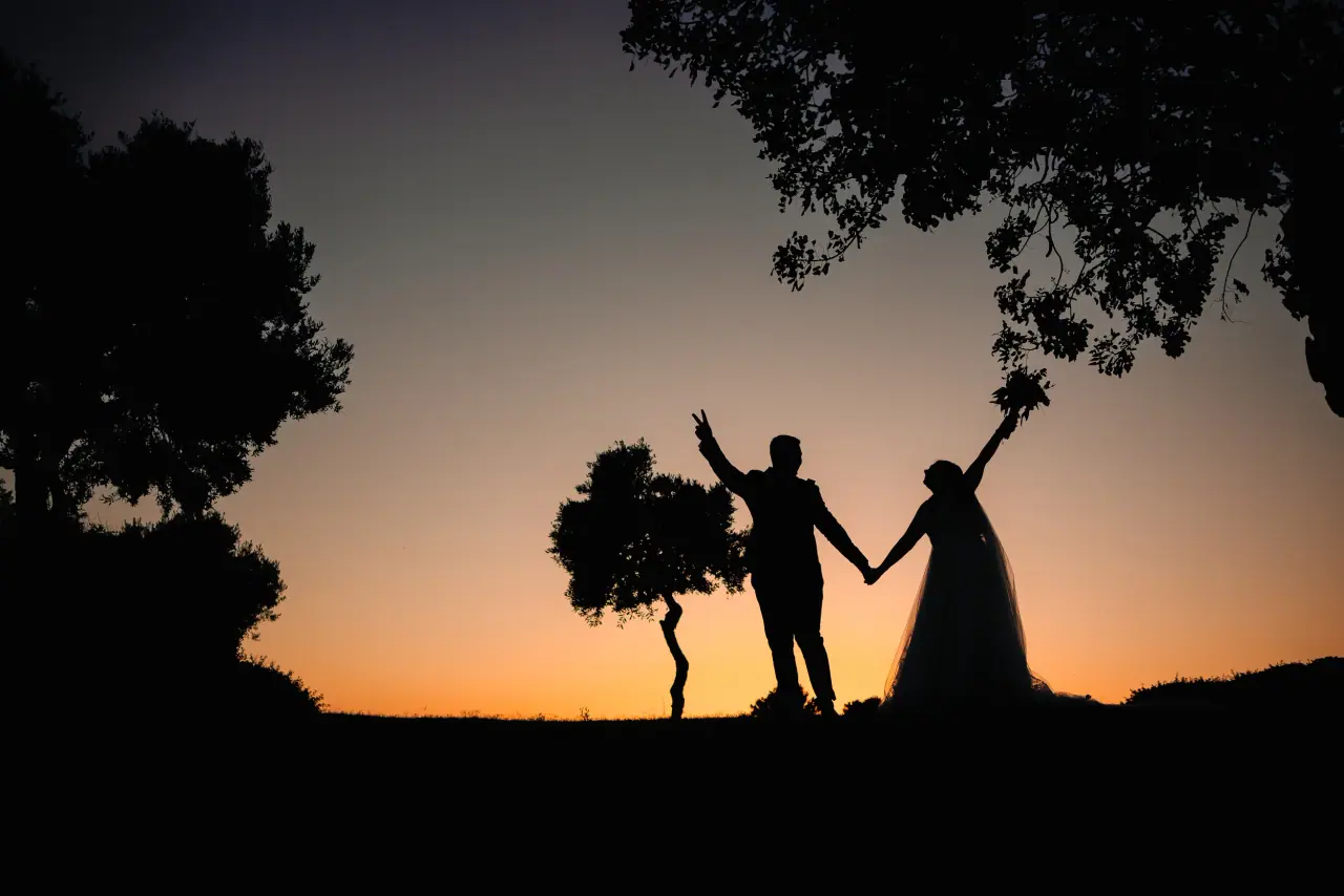 Ellis and Jonny silhouetted at sunset, hands and bouquet raised