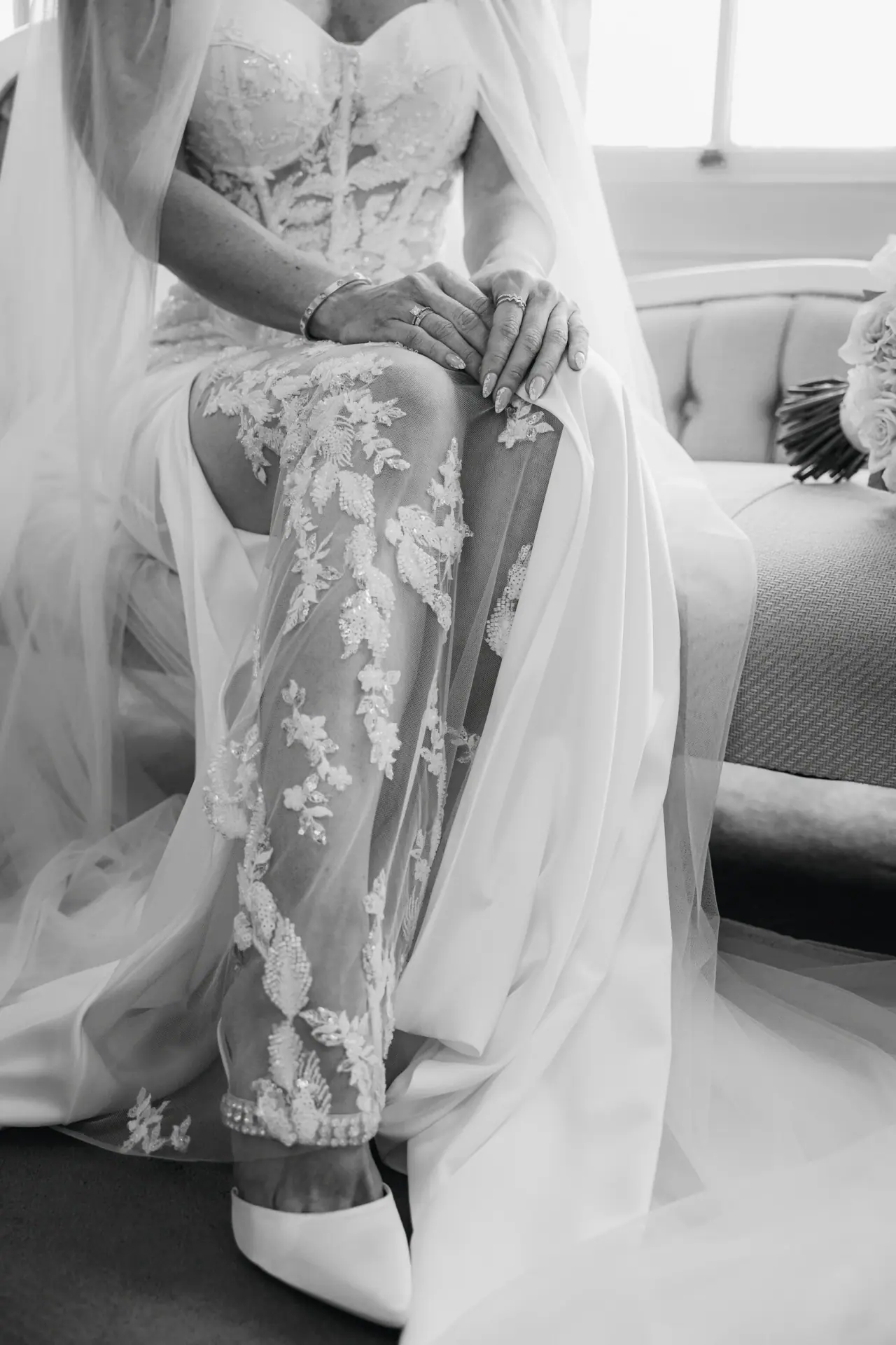 Black-and-white close-up of the bride's lace gown and shoes as she sits