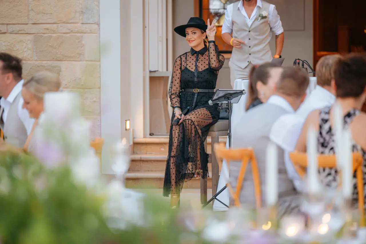 Singer in a black hat performing to guests seated at an outdoor reception, Anassa