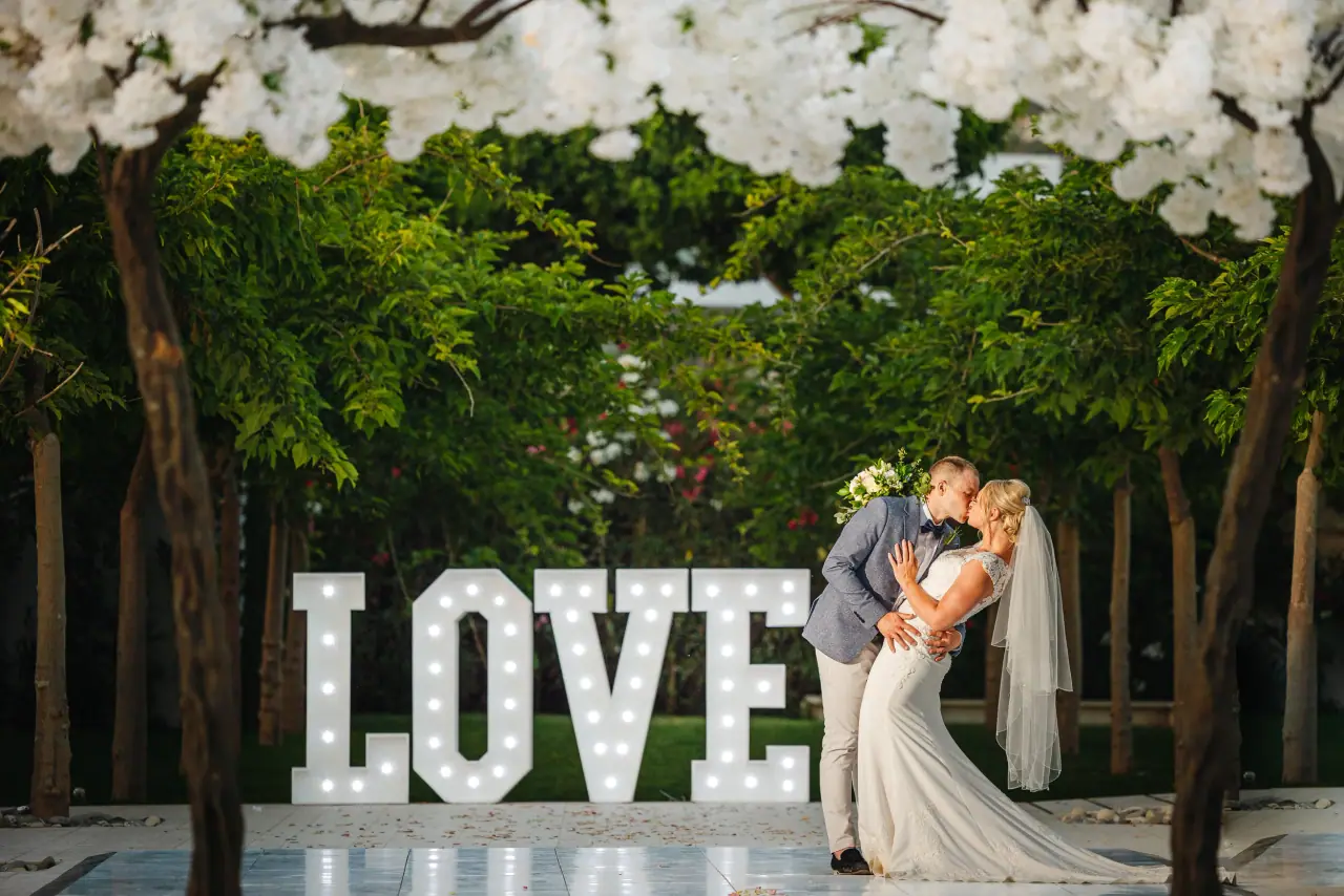 Jodie and Daniel kissing beside illuminated LOVE letters under a white floral canopy