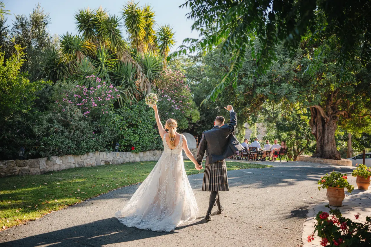 Samantha raising her bouquet as she walks with Dylan, in a kilt, at Vasilias Inn