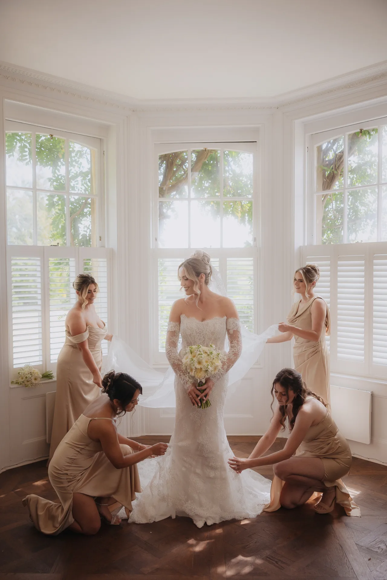 Bride surrounded by her bridesmaids as they help with her veil in a sunlit bay window at Northbrook Park, UK