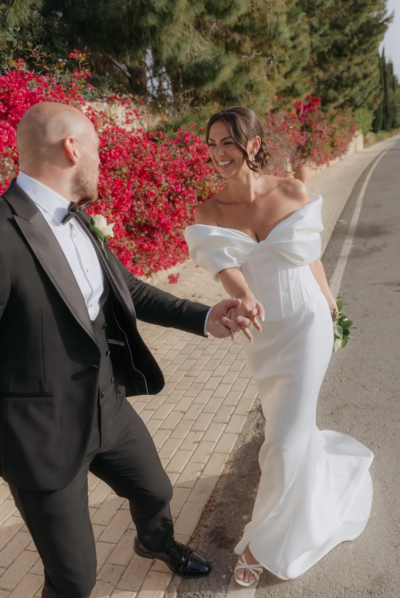 Bride and groom laughing hand in hand along a bougainvillea-lined road near Liopetro, Kouklia, Paphos