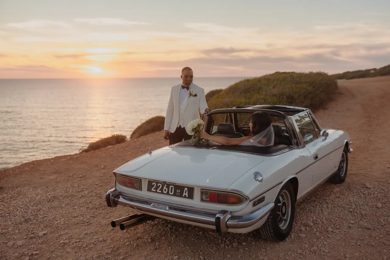 Groom standing beside the 1977 Triumph Stag as his bride sits inside, sunset over the sea in Cyprus