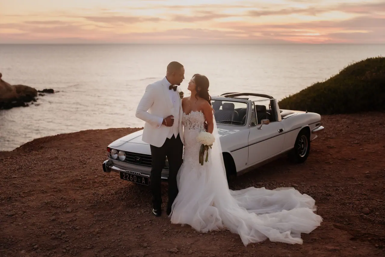 Bride and groom embracing beside the open-top Triumph Stag at sunset on the Cyprus coast