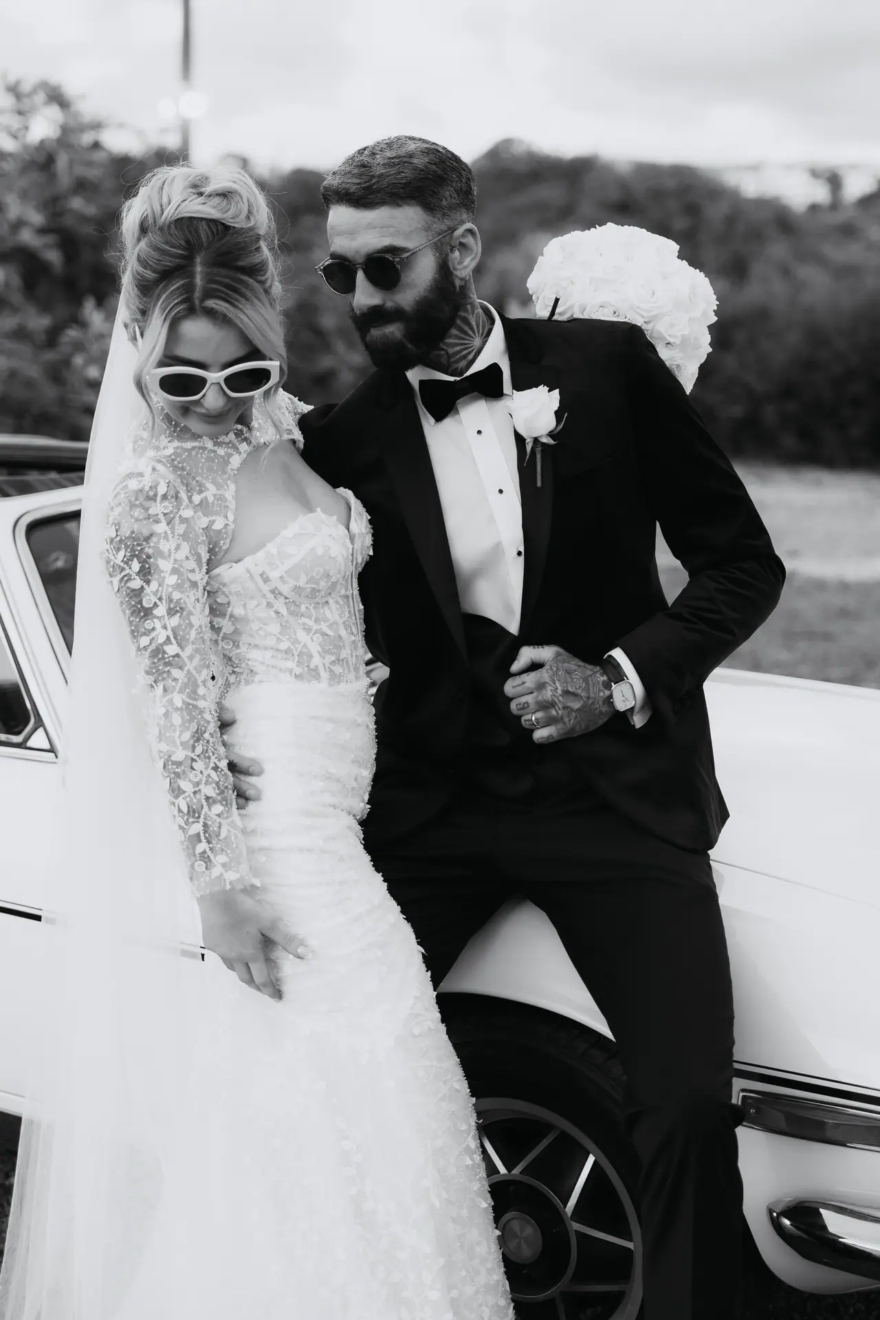 Black-and-white photo of the bride and groom in sunglasses leaning against the Triumph Stag